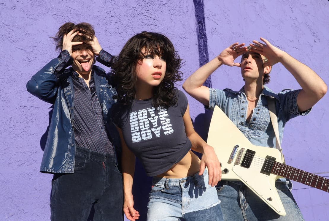 Three musicians posing with an electric guitar against a purple wall